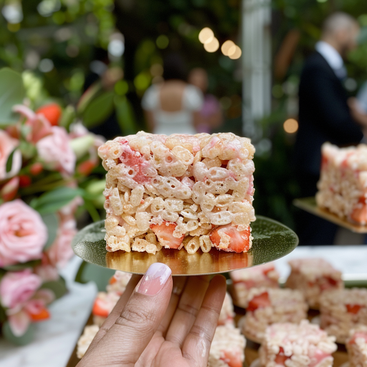  hand holds a slice of Strawberry Bliss rice crispy treat on a golden saucer, with a wedding reception in the background