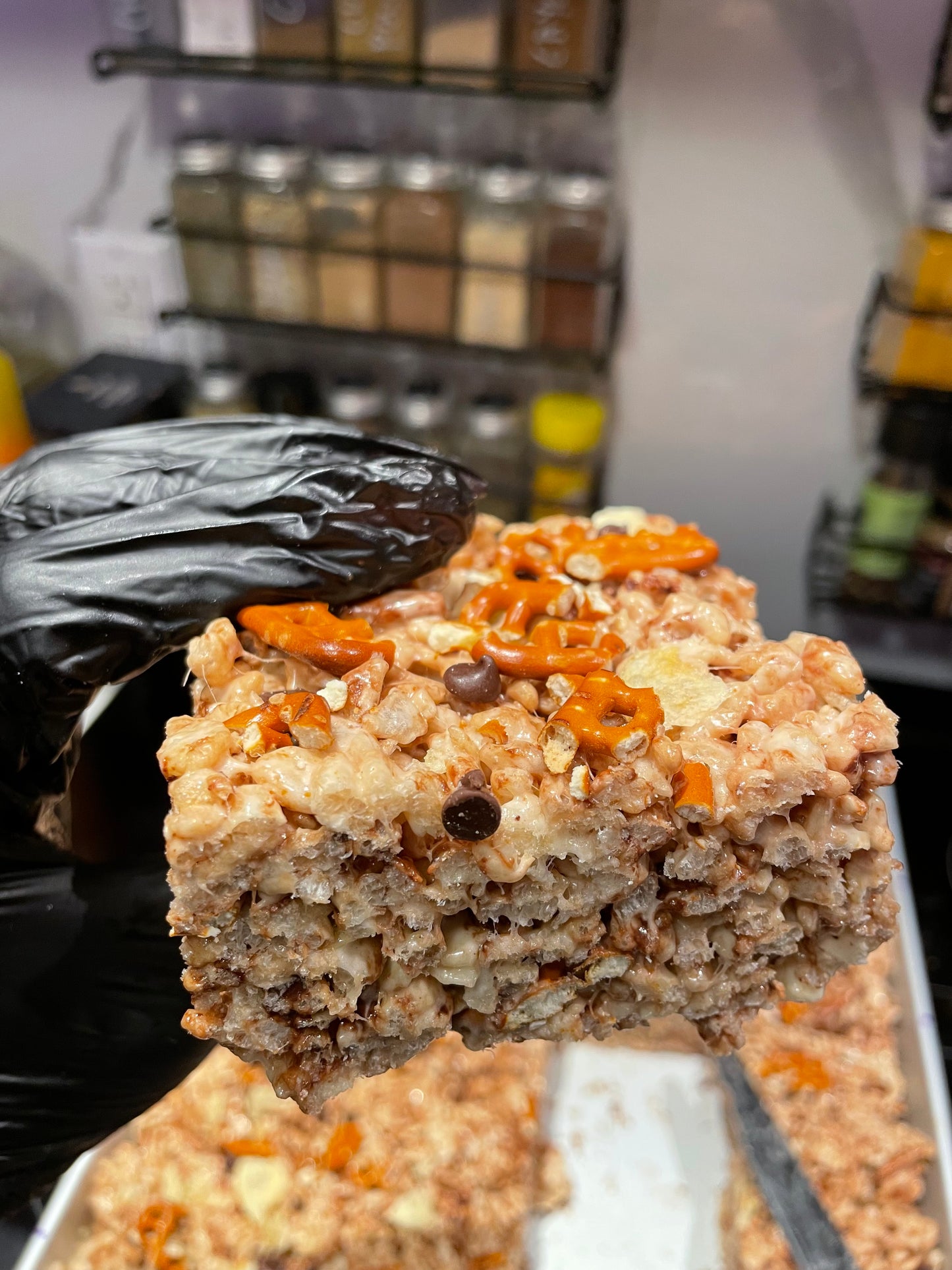 The image displays a close-up of a Rice Krispies treat held by a gloved hand, featuring a sprinkling of pretzels, potato chips, and chocolate chips on top. The background is blurred but suggests a kitchen or cafe setting with jars of ingredients.