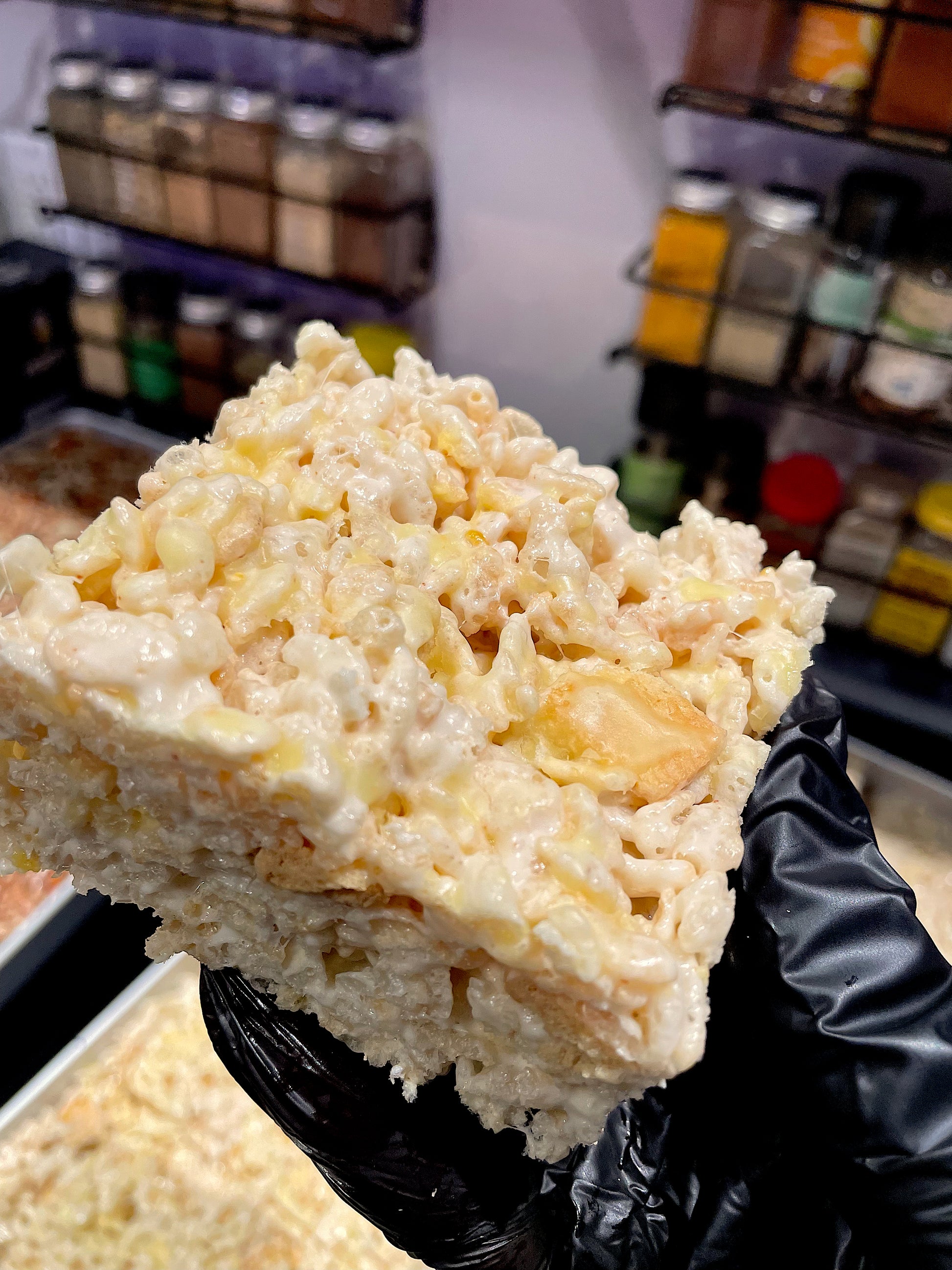 The image shows a close-up of a Rice Krispies treat with lemon cookie chunks and a drizzle of lemon icing, held in a gloved hand. The treat looks rich and zesty, and the background suggests a kitchen setting.