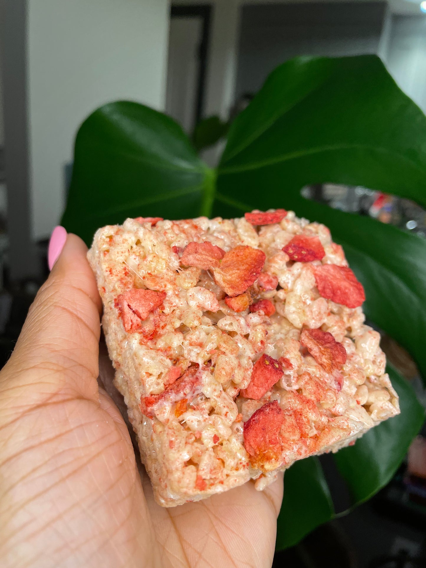 The image features a hand holding a Rice Krispies treat with strawberry pieces, set against a natural green leaf backdrop, indicating a fresh, homemade quality.