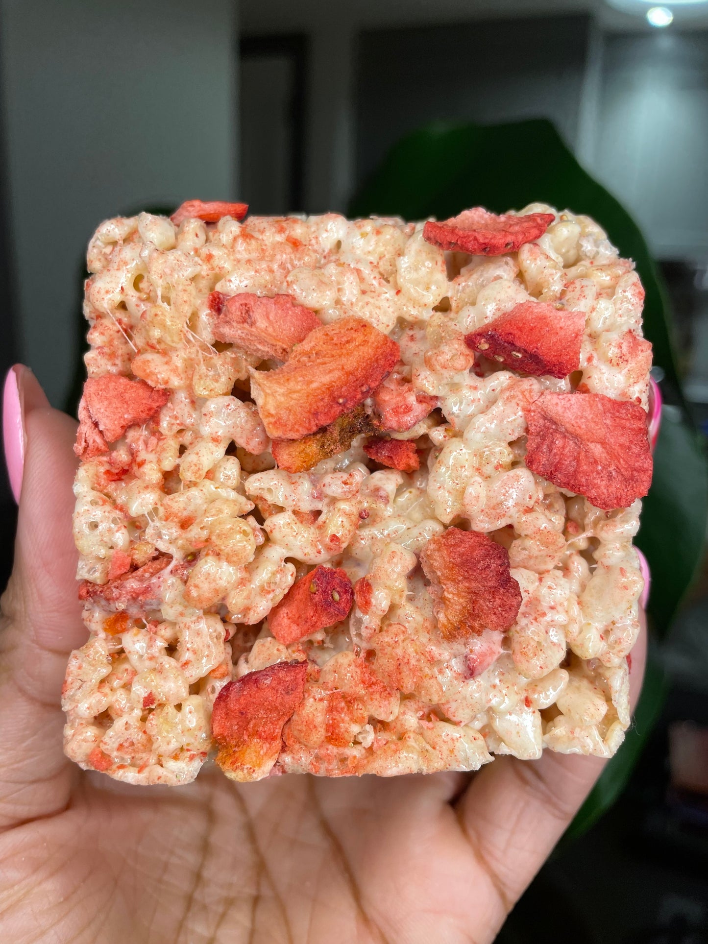 The image features a hand holding a Rice Krispies treat with strawberry pieces, set against a natural green leaf backdrop, indicating a fresh, homemade quality.   User Uploaded image ChatGPT The image shows a close-up of a hand holding a square Rice Krispies treat speckled with vibrant red strawberry pieces, with a lush green leaf providing a contrasting background.