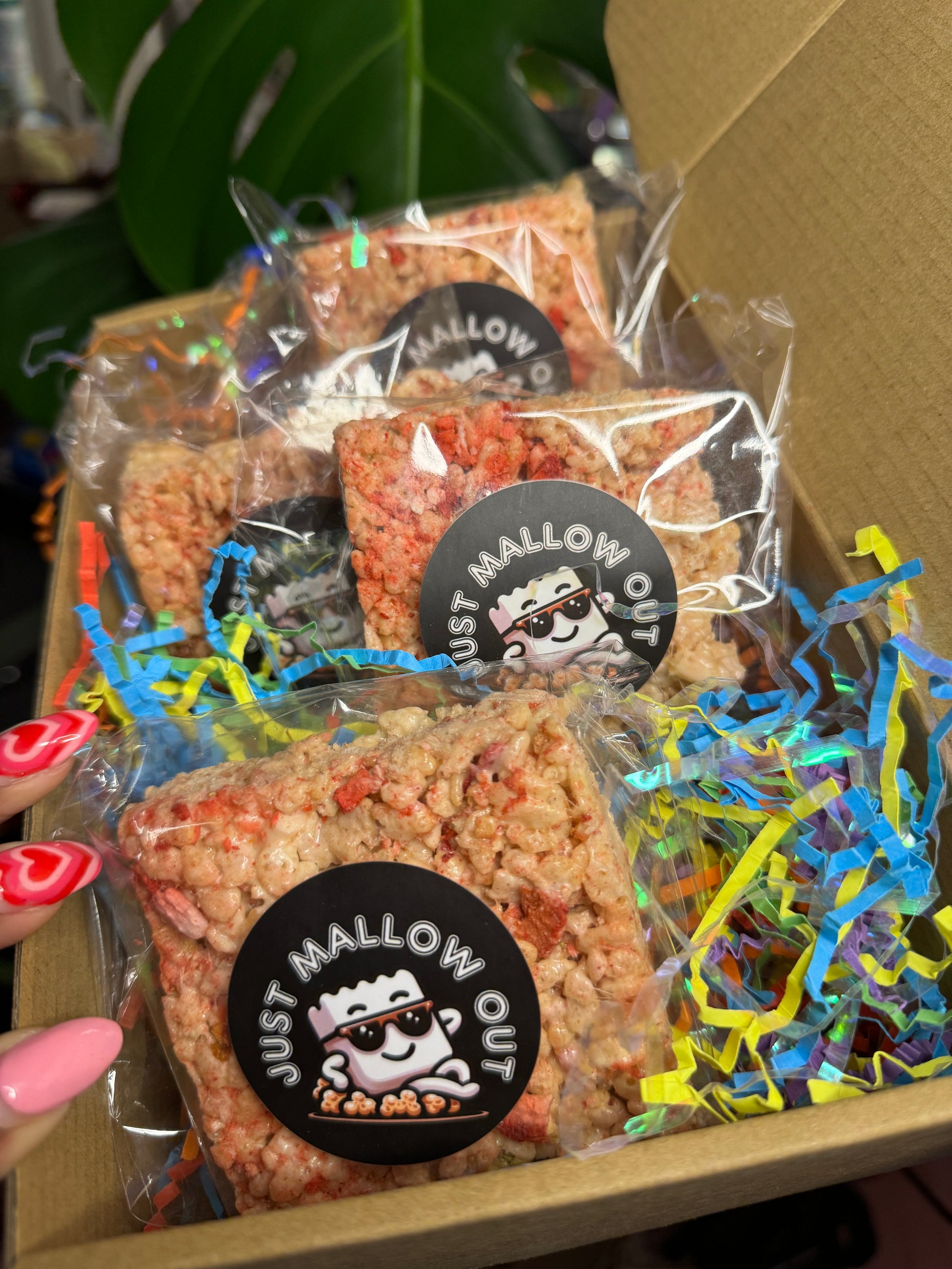 The image depicts individually wrapped Strawberry Bliss Rice Krispies treats inside a box, ready for distribution. Each treat is sealed in clear plastic with a "JUST MALLOW OUT" sticker featuring a smiling marshmallow chef logo. The box is accented with colorful shredded paper, creating a festive and inviting presentation.